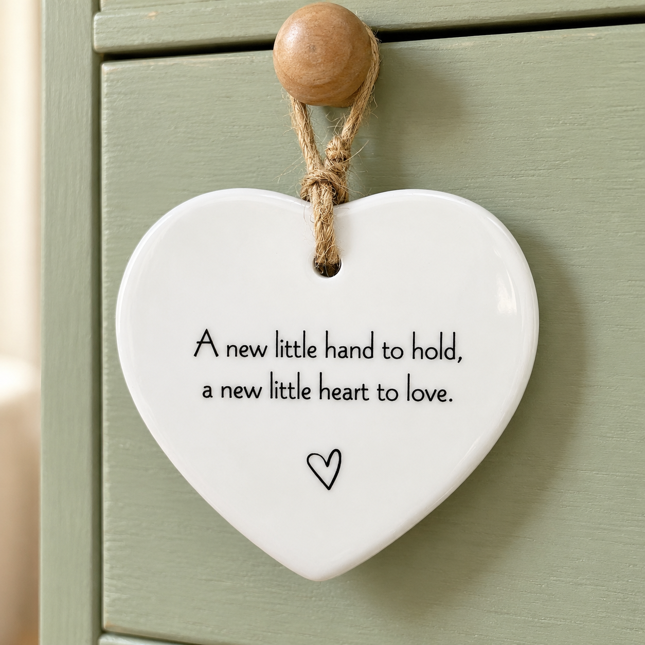 New baby ceramic heart keepsake with the words A new little hand to hold, a new little heart to love, hanging on a sage green cupboard