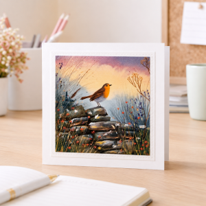 Handmade robin greeting card standing upright on a wooden desk alongside a notebook, pen, and dried flowers.