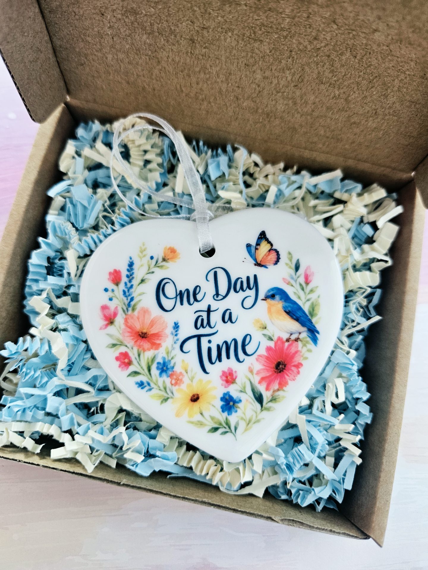 One Day at a Time ceramic heart keepsake nestled inside an open kraft gift box filled with light blue and cream shredded paper filler.