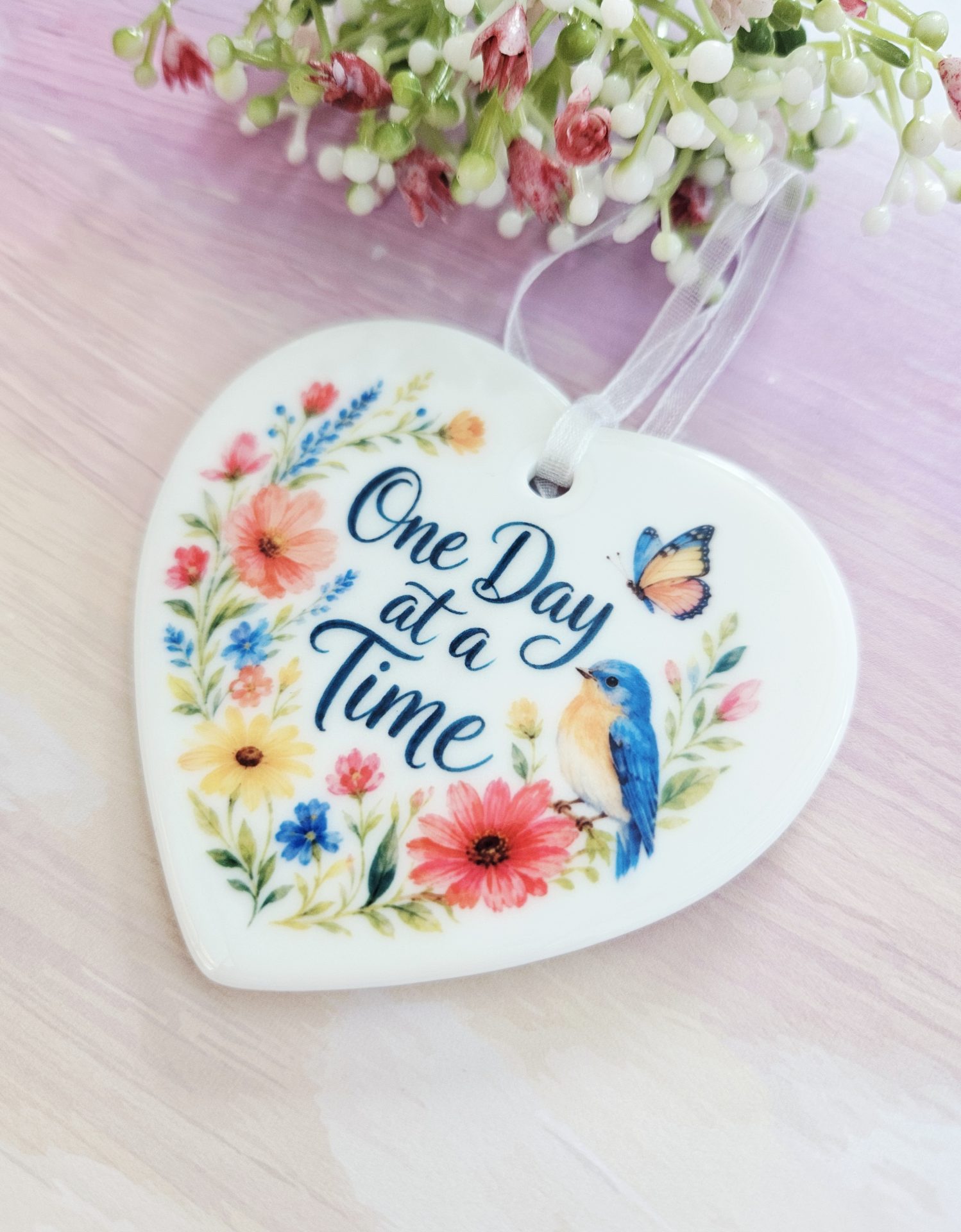 Flat lay product shot of the One Day at a Time ceramic heart keepsake resting on a light wood surface with decorative white and pink florals at the top.