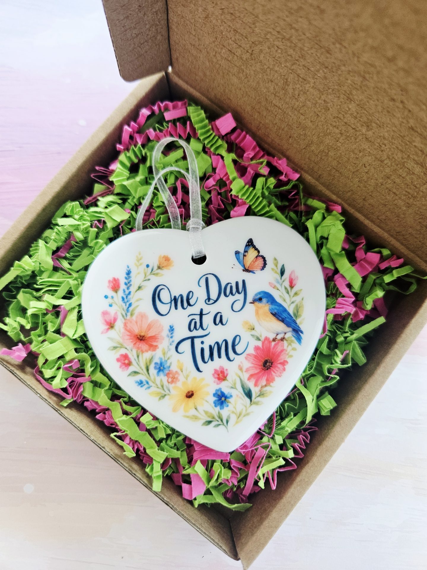 One Day at a Time ceramic heart keepsake nestled in an open kraft gift box filled with vibrant green and pink shredded paper filler.