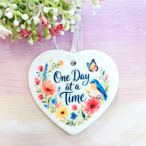 One Day at a Time ceramic heart keepsake with wildflower illustration, blue bird, butterfly, and white ribbon against a pink background.
