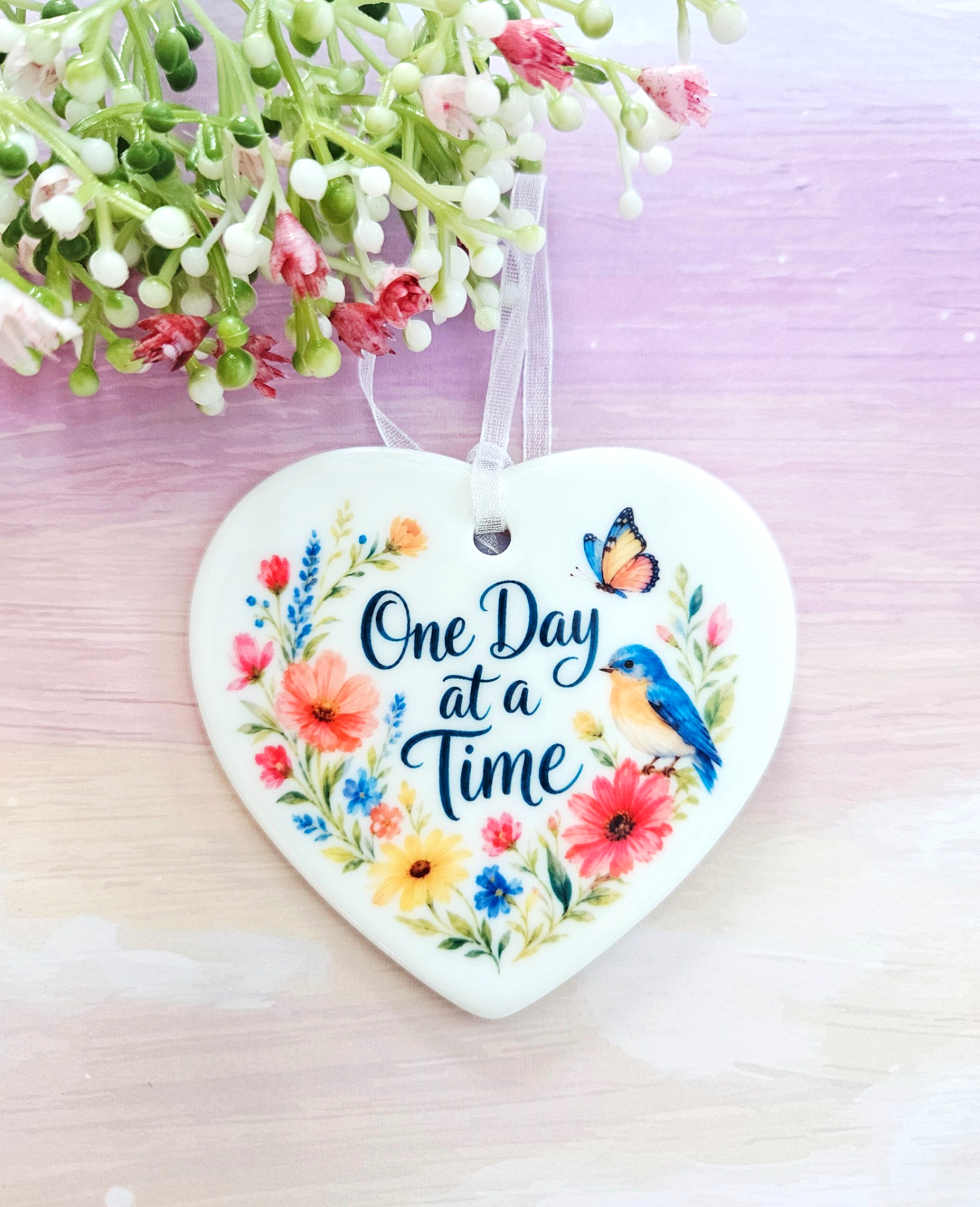 One Day at a Time ceramic heart keepsake with wildflower illustration, blue bird, butterfly, and white ribbon against a pink background.