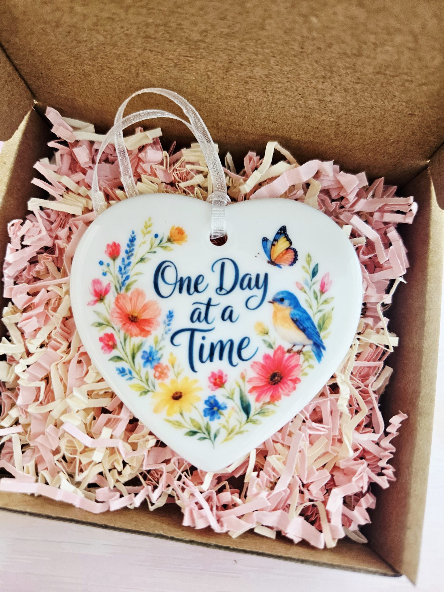 One Day at a Time ceramic heart keepsake displayed in an open kraft gift box filled with pink and cream shredded paper filler.