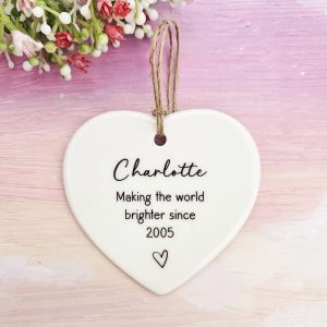 Personalised 21st birthday ceramic heart keepsake featuring the name Charlotte and the text Making the world brighter since 2005 with a small heart motif, hung with twine against a pink and white background with small floral details.